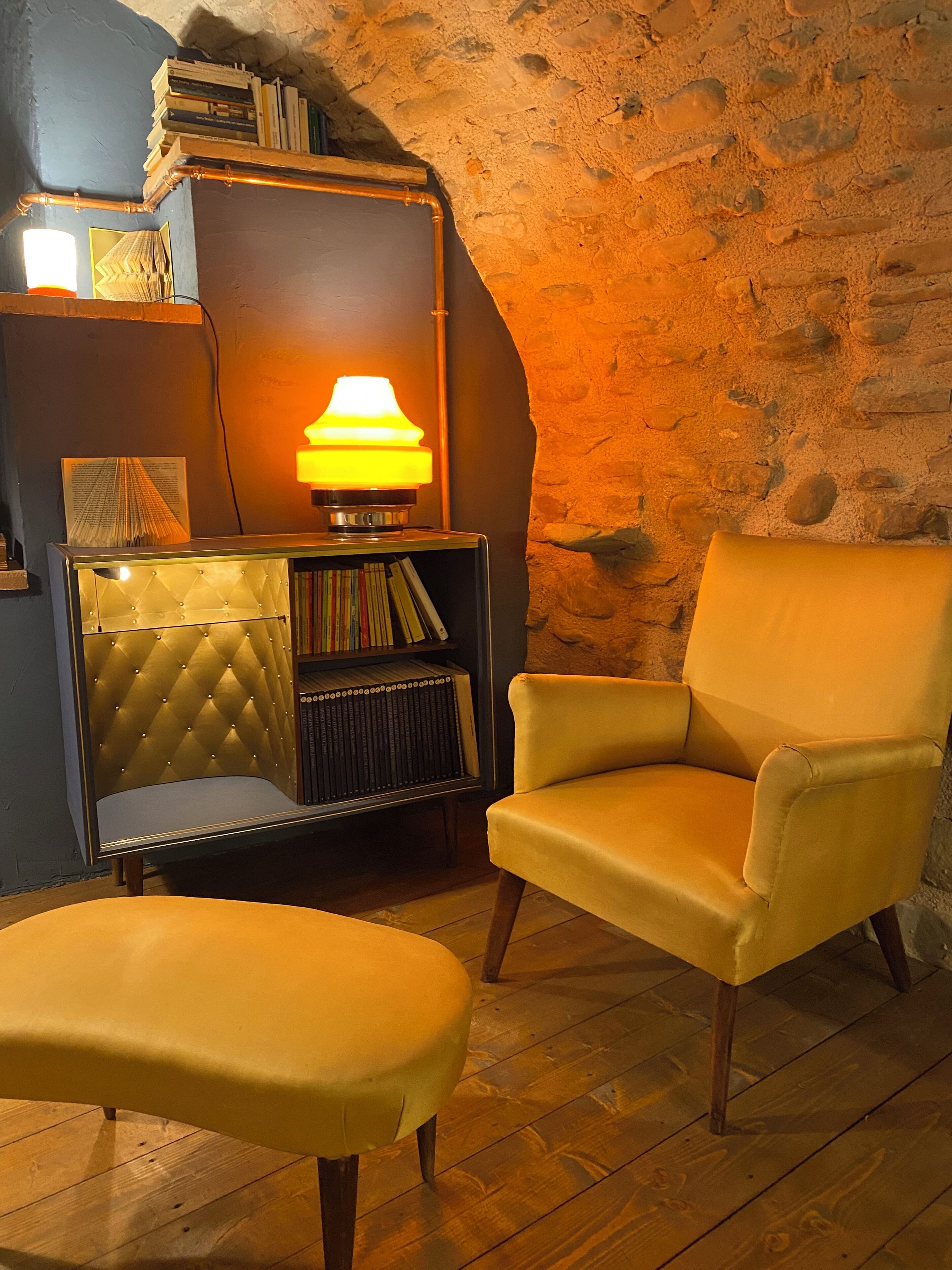 Una habitación acogedora con pared de piedra, una estantería, una lámpara y un sillón amarillo.