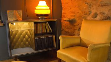 Una habitación acogedora con pared de piedra, una estantería, una lámpara y un sillón amarillo.