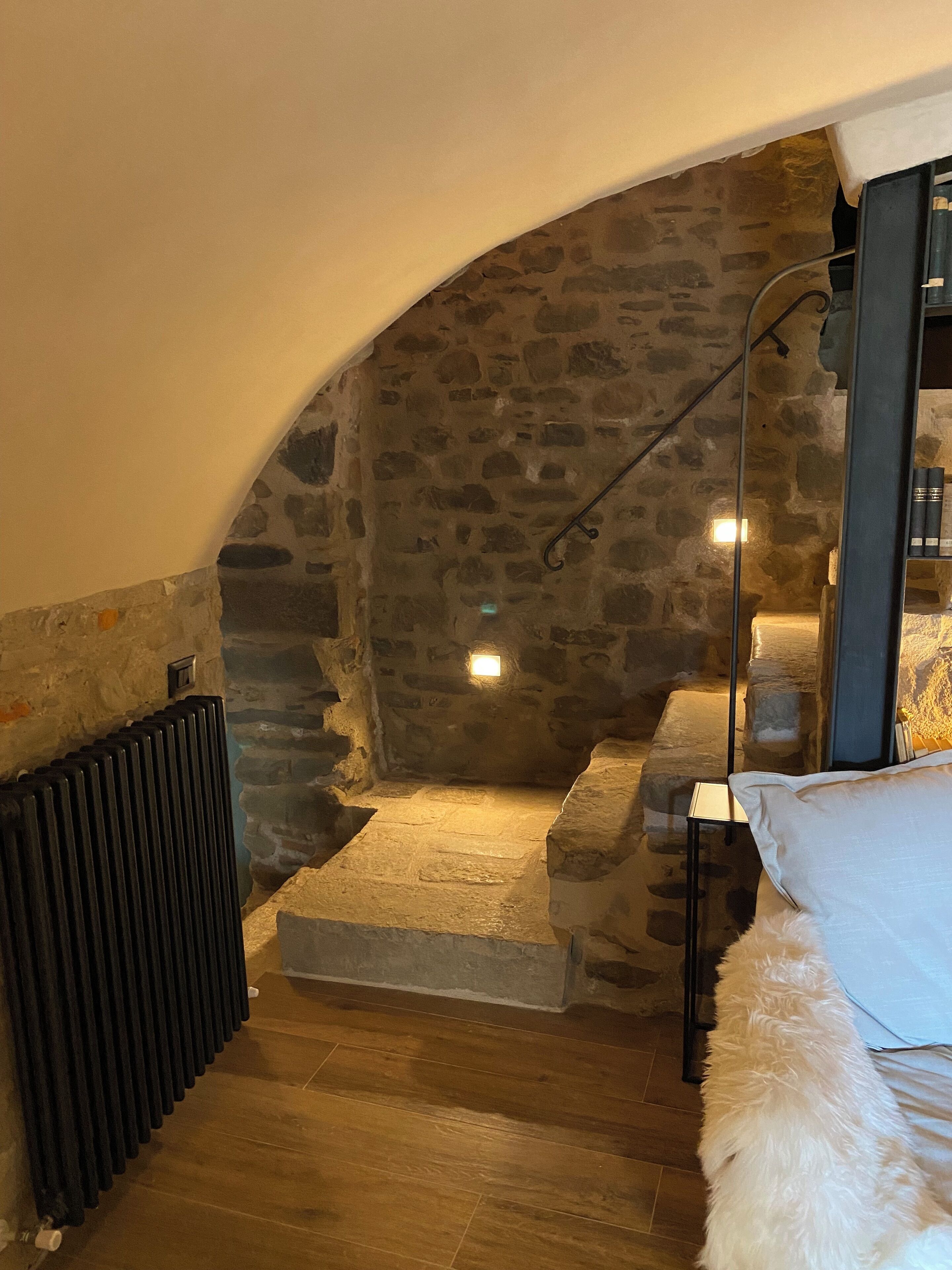 A cozy attic bedroom with stone walls, a wooden floor, a bed with white bedding, and a small table with books.