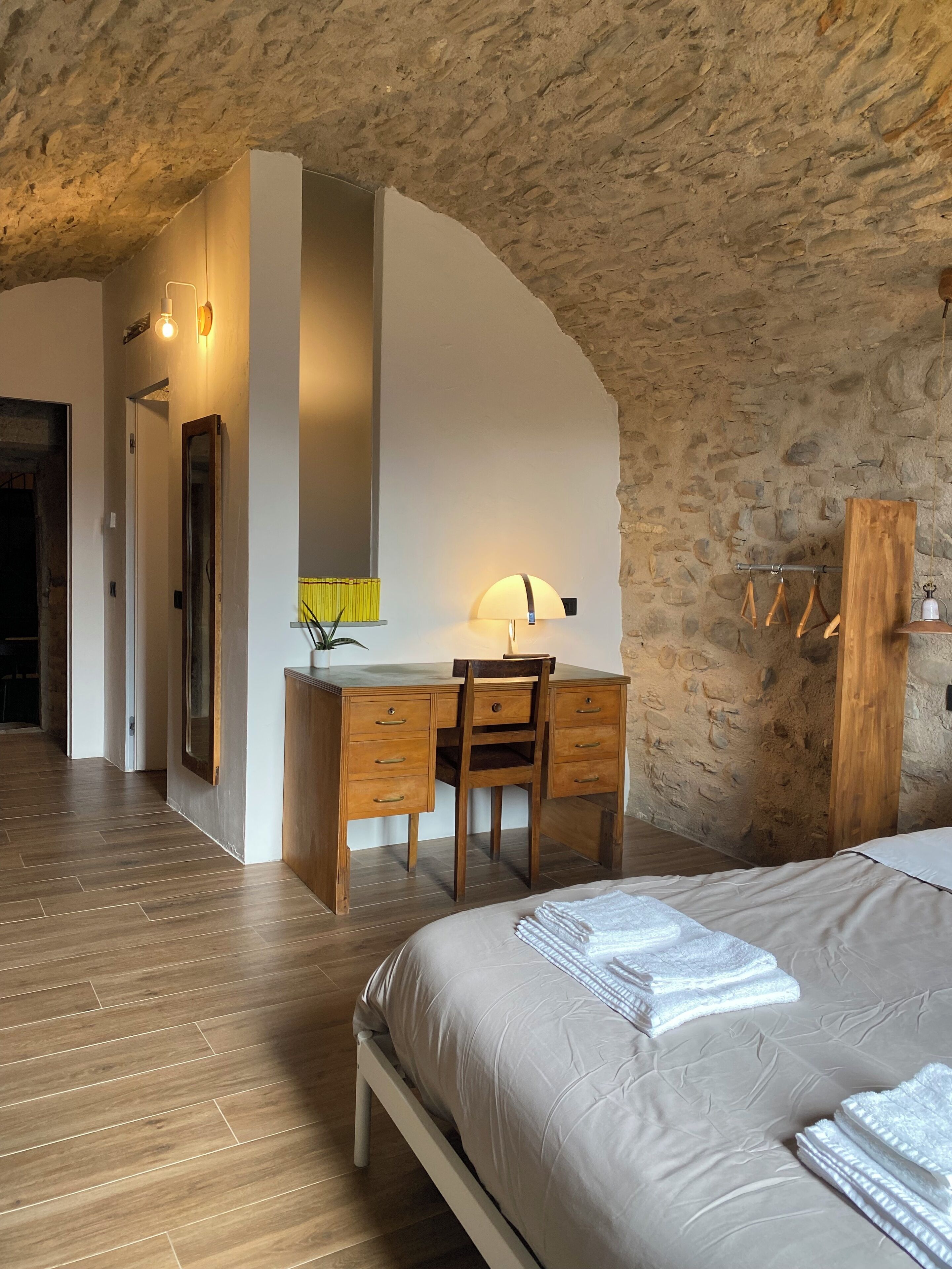 A bedroom with a wooden desk, a chair, a bed with neatly folded towels, and a stone wall.
