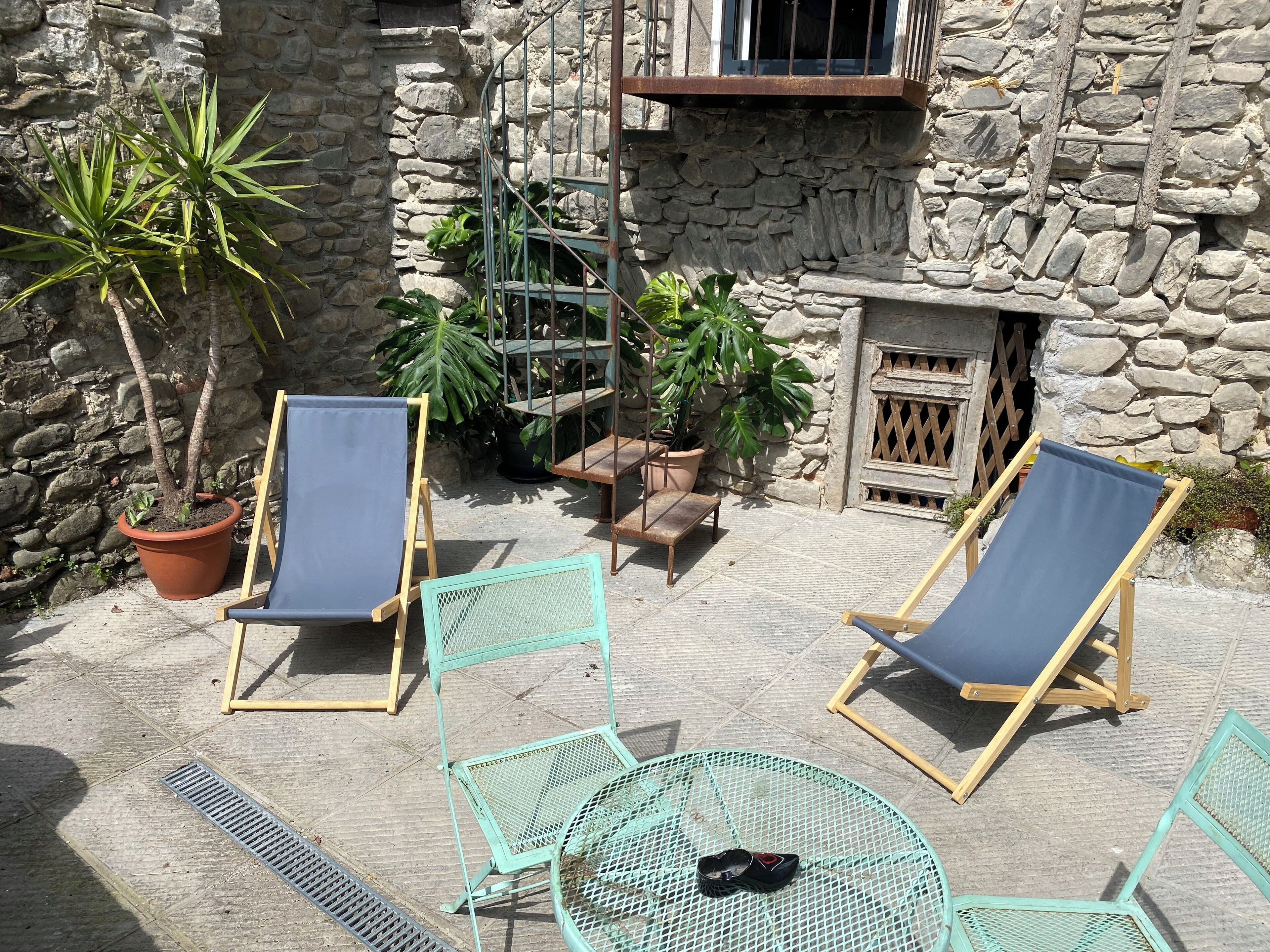 A stone building with a small patio area featuring two blue lounge chairs, a round table, and potted plants.