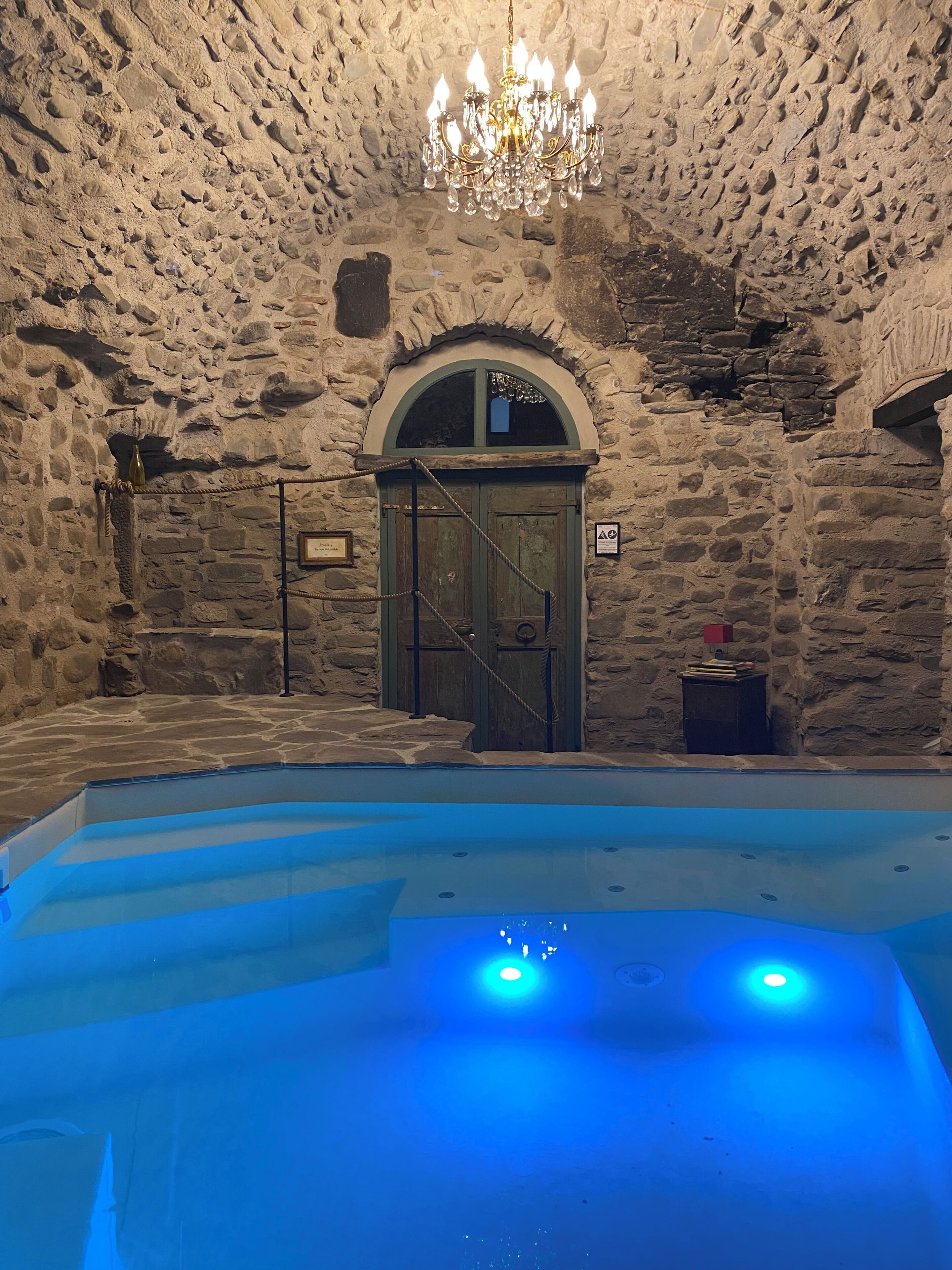 Una stanza con pareti in pietra, una piscina, un lampadario e una porta in legno.