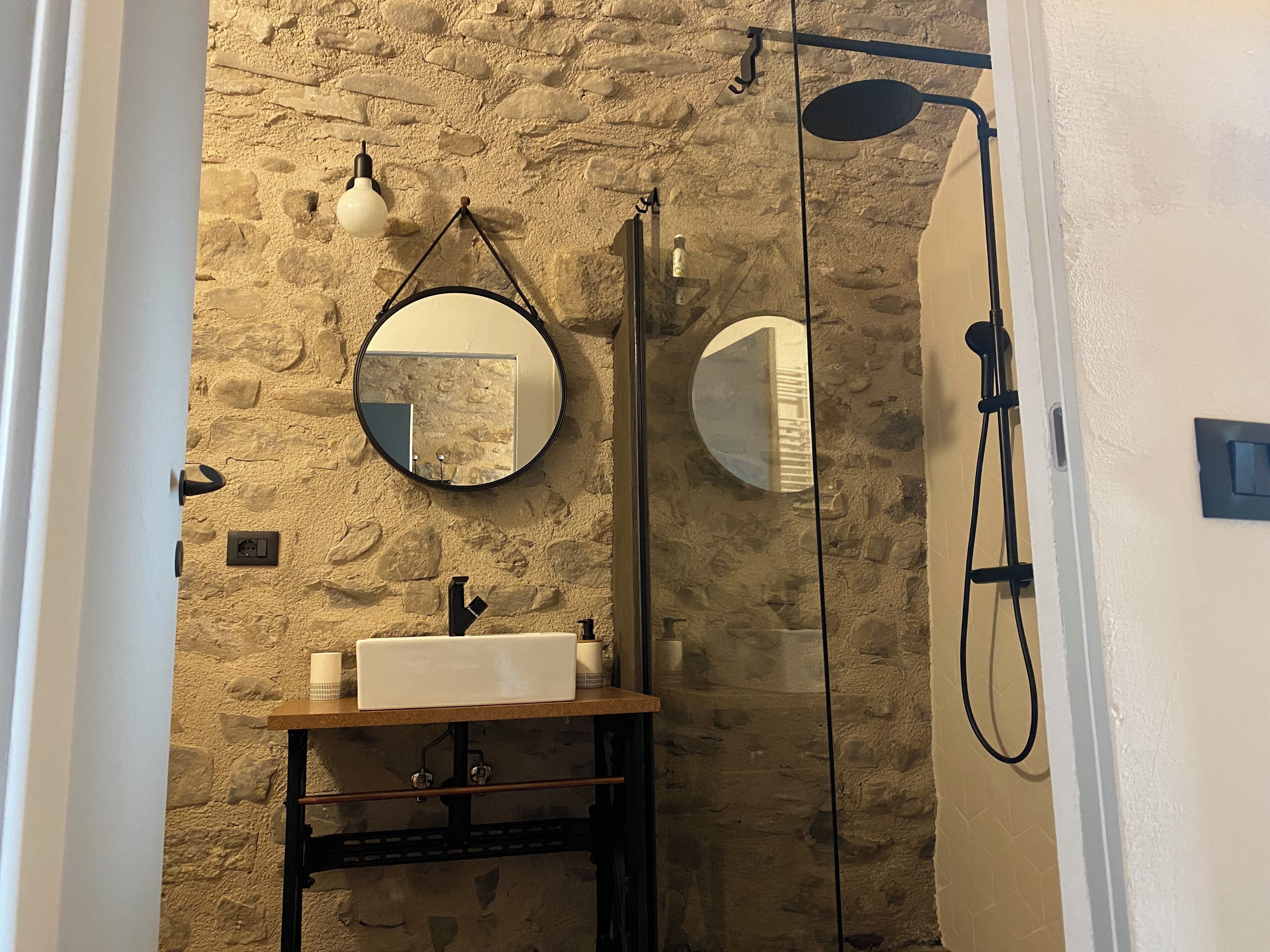 A bathroom with a stone wall, a round mirror, a sink, and a shower.