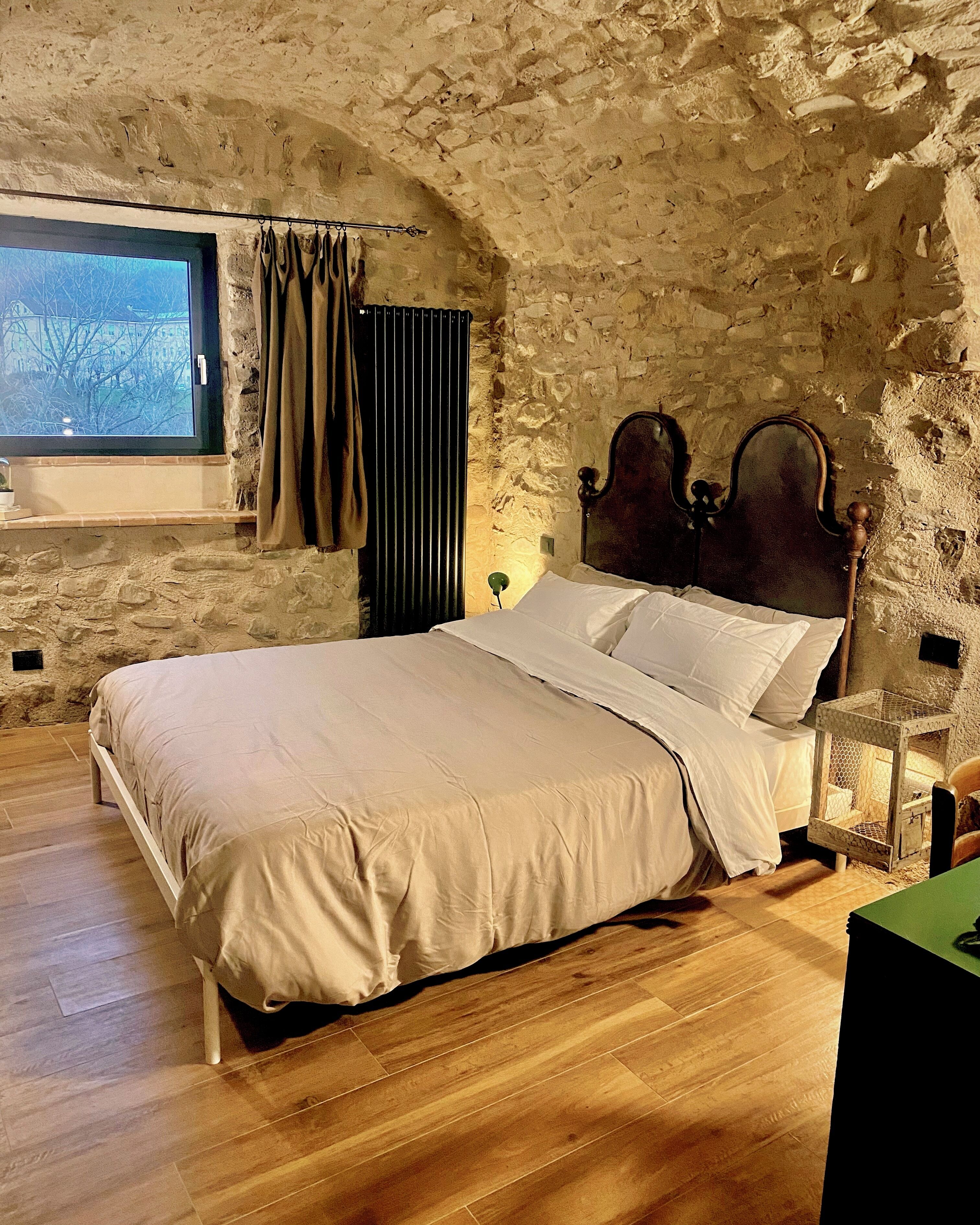 Une chambre avec un mur en pierre, un lit en bois, une fenêtre avec des rideaux et une table de chevet avec une lampe.