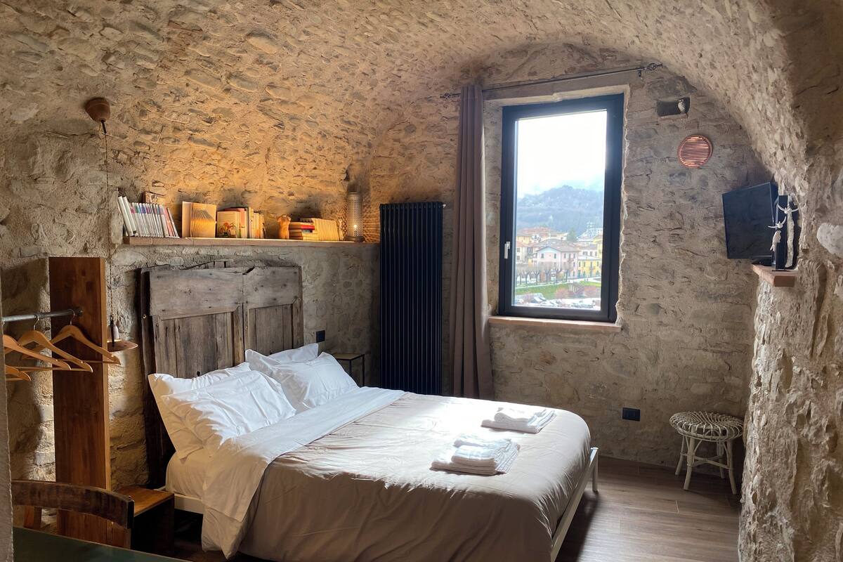 Een rustieke slaapkamer met een stenen muur, een houten bed, een klein tafeltje en een raam met uitzicht op gebouwen.