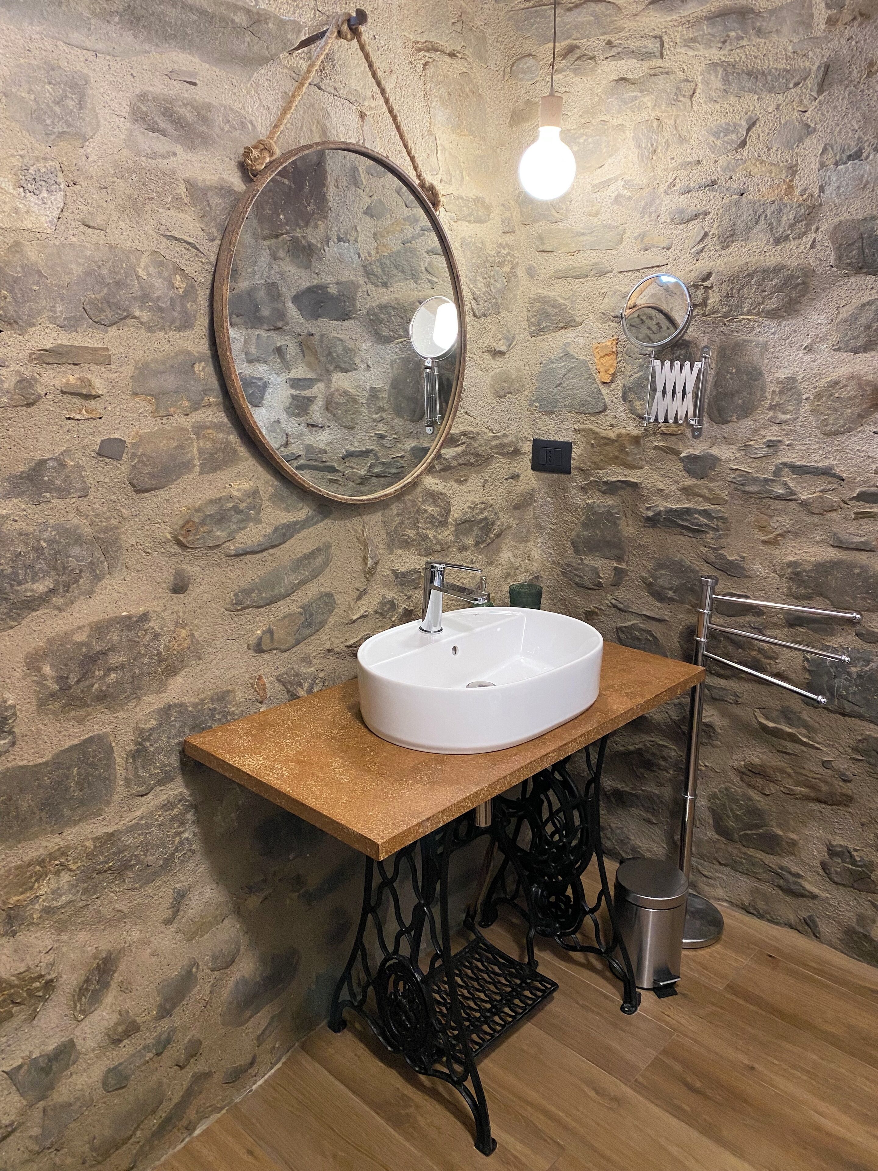 A bathroom with a stone wall, a round mirror, a white sink, a wooden countertop, a metal faucet, a hanging light bulb, a towel rack, and a trash bin.