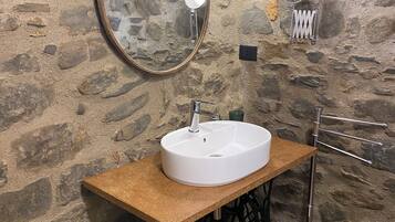 A bathroom with a stone wall, a round mirror, a white sink, a wooden countertop, a metal faucet, a hanging light bulb, a towel rack, and a trash bin.