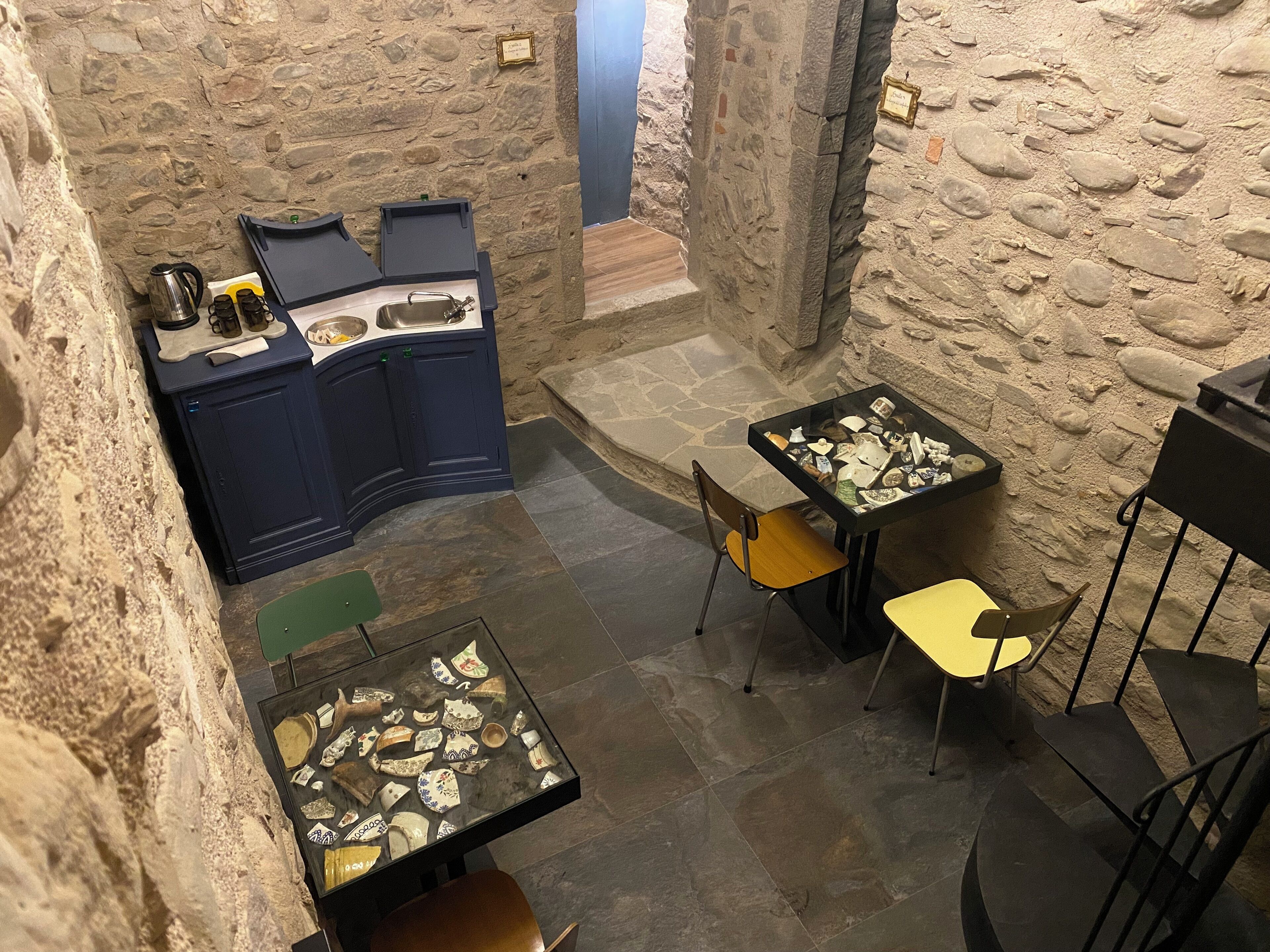 Una habitación con paredes de piedra, una kitchenette, dos mesas con sillas y una pequeña escalera.
