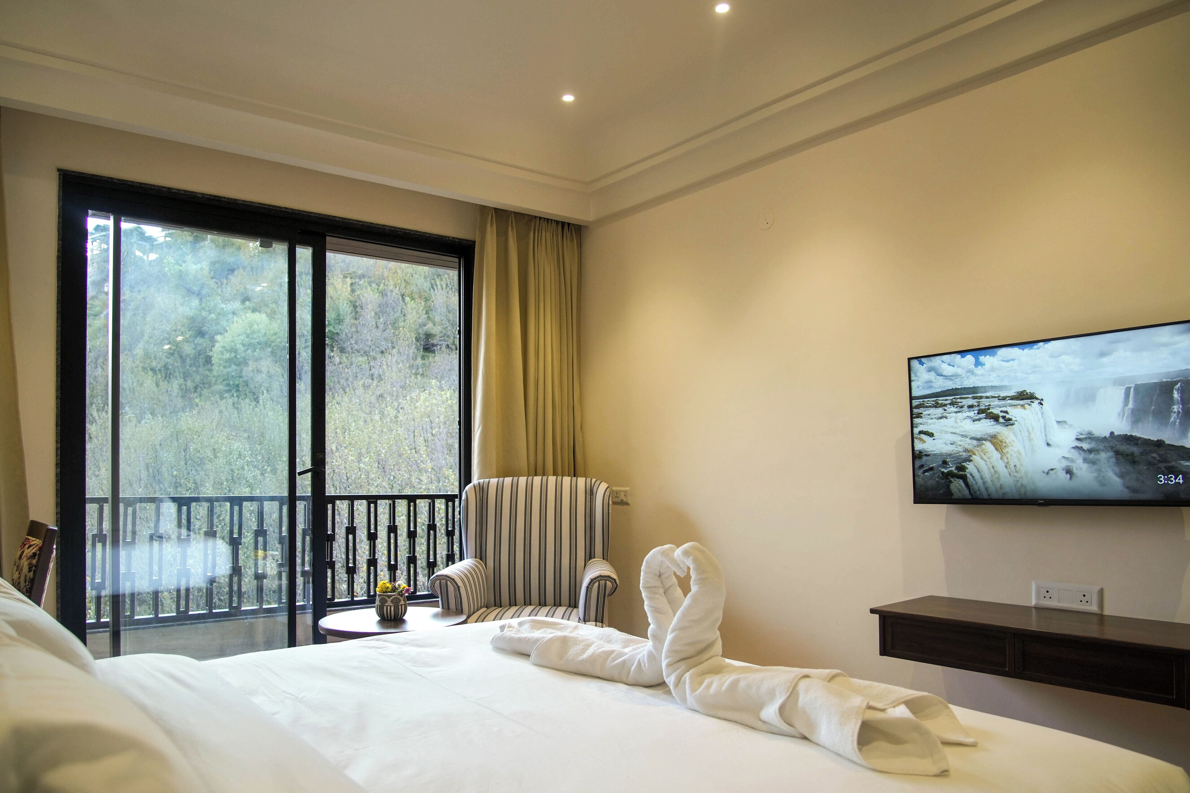 A hotel room with a large bed, a TV on the wall, a chair, and a balcony with a view of trees.