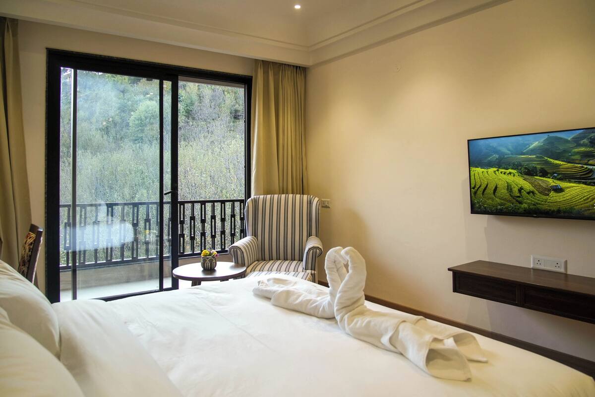 A hotel room with a large bed, a TV mounted on the wall, a balcony with a view of greenery, and a striped armchair.