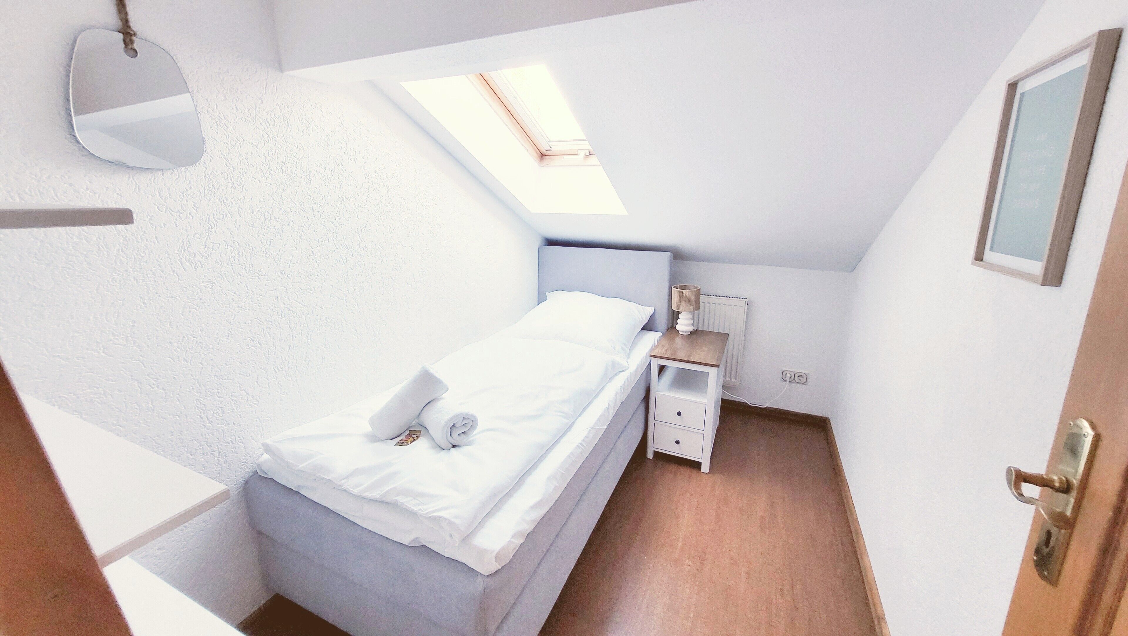 Un petit lit bien fait, placé dans un coin de la pièce, avec un puits de lumière au plafond, une table de chevet avec une lampe et une petite commode.