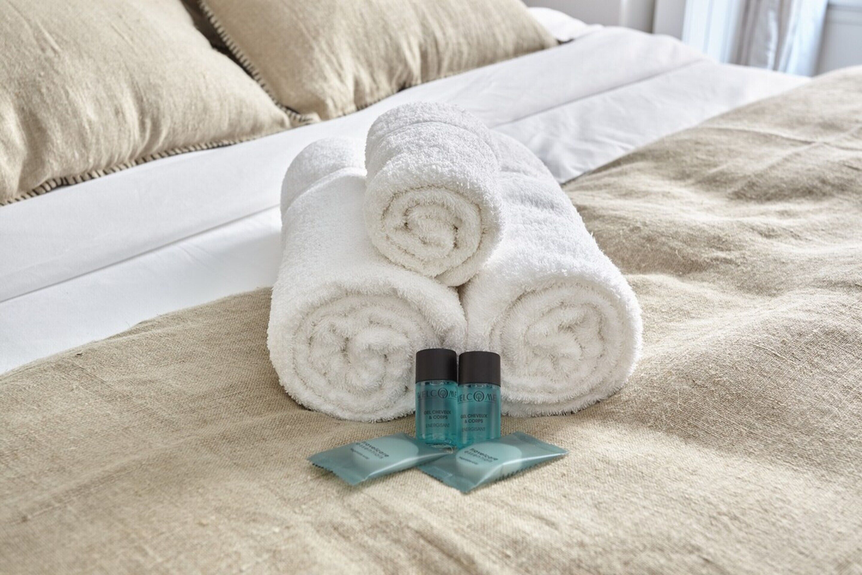 Una cama bien hecha con toallas blancas enrolladas y dos botellas pequeñas sobre un edredón color beige.