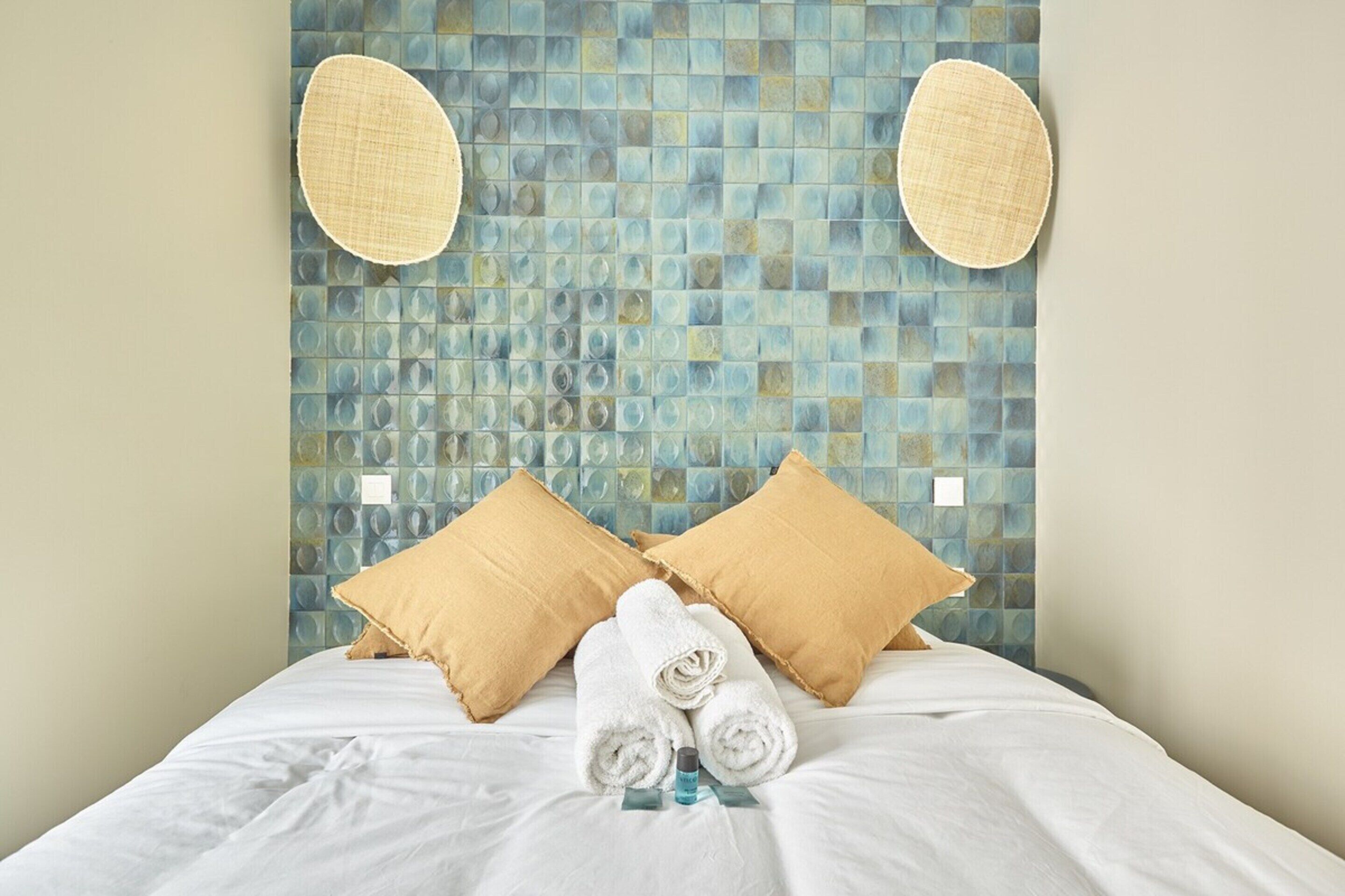 A neatly made bed with rolled white towels and two beige pillows, a blue mosaic wall behind, and a small blue bottle on the bed.