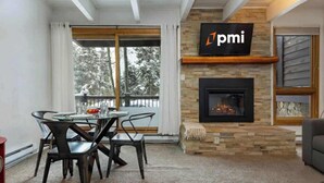 A modern living room with a fireplace, a dining area with a glass table and chairs, and a TV mounted on the wall.