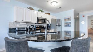 A modern kitchen with white cabinets, stainless steel appliances, and a glass island with two chairs. - Ocean Hibiscus Retreat – For 8 (St. Augustine)