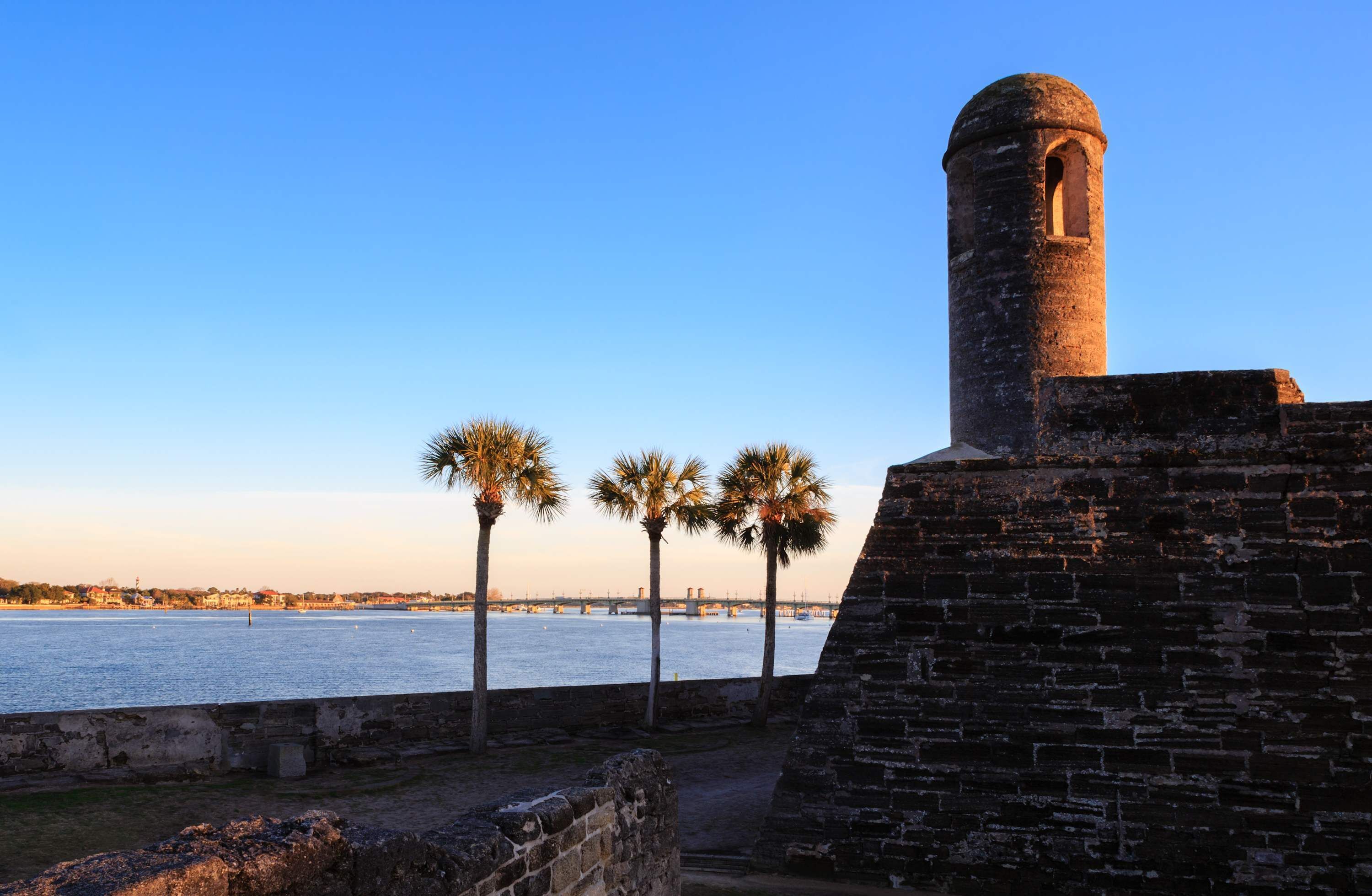 A historic fort with a brick tower overlooking a calm body of water, palm trees in the foreground, and a distant bridge.