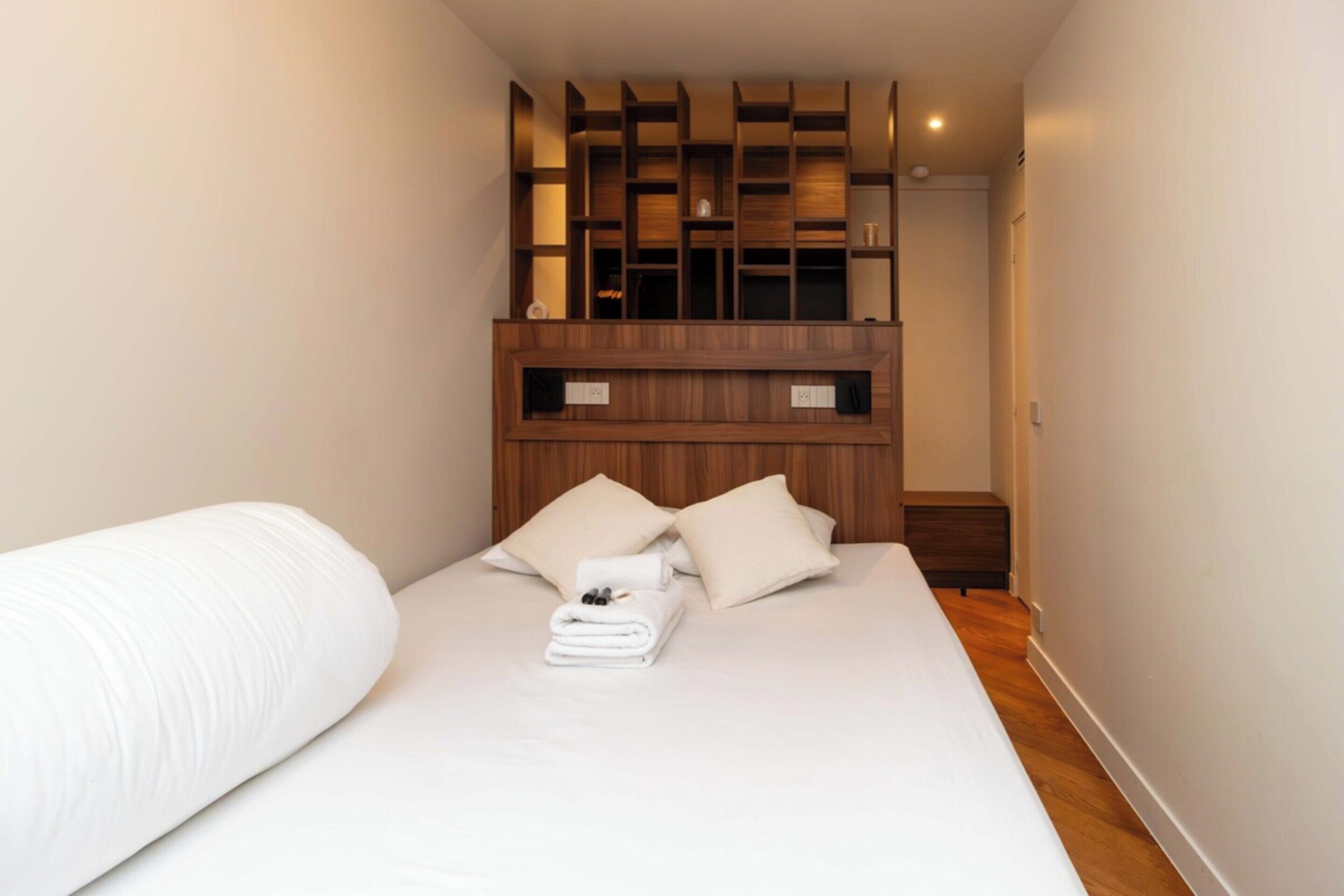 A neatly made bed with a white comforter and pillows, a wooden headboard, and a small wooden nightstand with a folded towel.