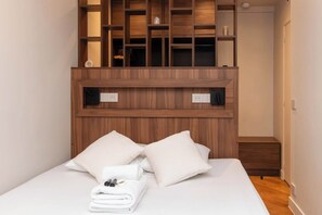 Un lit bien fait, avec des oreillers et une serviette pliée, une tête de lit en bois dotée d’étagères intégrées et une petite table de chevet en bois.