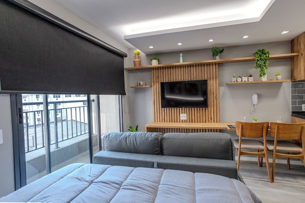 A modern bedroom with a large bed, a TV mounted on a wooden wall, a desk with a chair, and a balcony with a view of buildings.