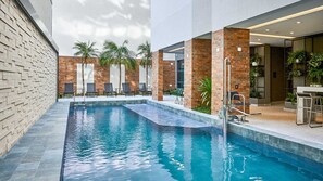 A swimming pool with a brick wall on one side and a glass wall on the other. There are lounge chairs and a bar area with plants.