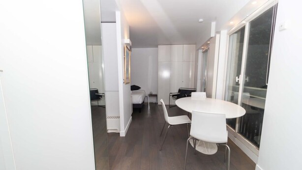A modern, well-lit room with a round dining table and white chairs, a bed with a black headboard, and a large sliding glass door leading to a balcony. - Charming Studio Paris / Le Marais (Paris)