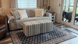 Una habitación con sofá, reposapiés, alfombra estampada, ventilador de techo y vistas a través de un ventanal.
