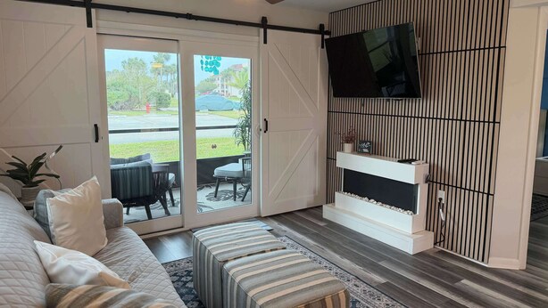 Una sala de estar con un sofá, otomanas, un televisor montado en la pared y un ventanal corredizo que da a un área exterior.