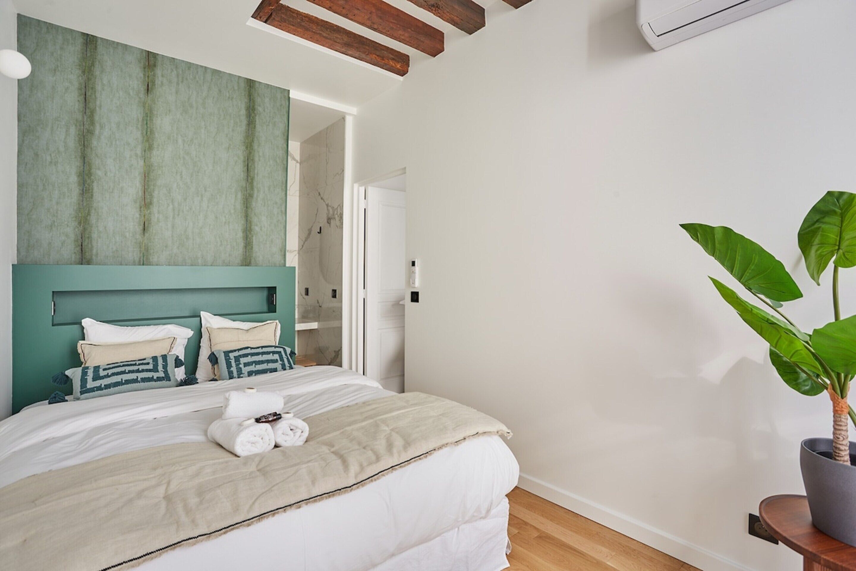 A bedroom with a bed, a headboard, a potted plant, and a door leading to another room.