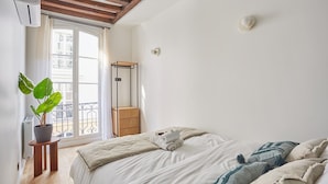 Ein Schlafzimmer mit Bett, einer Topfpflanze, einem Nachttisch aus Holz und einem Fenster mit Blick nach draußen.