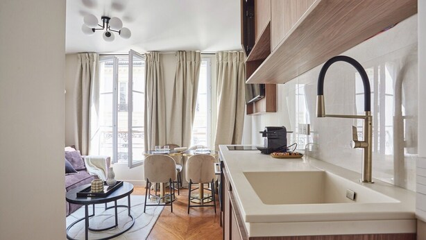A modern kitchen with a large island, a built-in oven, and a stainless steel faucet. There is a dining area with a round table and chairs, and a sofa in the background. - New Apartment Center of Paris/le Marais (Paris)