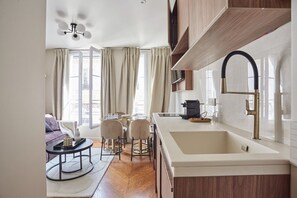 A modern kitchen with a large island, a built-in oven, and a stainless steel faucet. There is a dining area with a round table and chairs, and a sofa in the background. - New Apartment Center of Paris/le Marais (Paris)