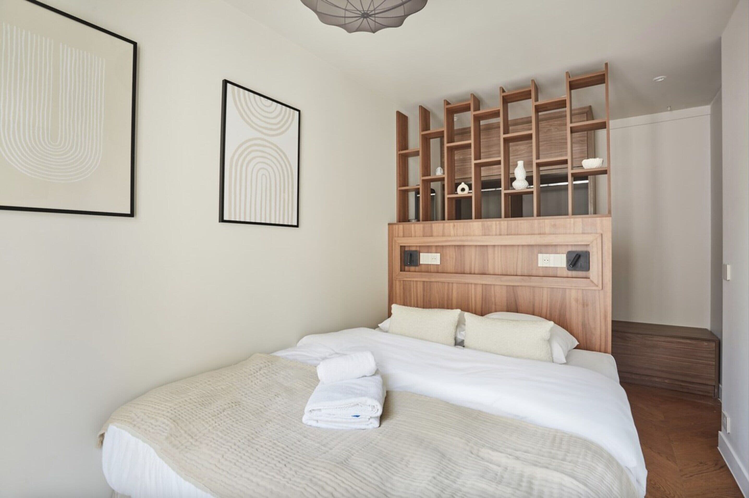 A neatly made bed with white linens and towels, a wooden headboard with shelves, and two framed artworks on the wall.