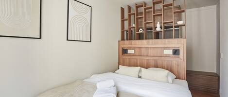 A neatly made bed with white linens and towels, a wooden headboard with shelves, and two framed artworks on the wall.