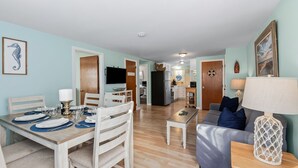 A spacious living area with a dining table set for four, a sofa, and a kitchen in the background. - Walk to Beach, Steps from Park, Pet-Friendly & Fire Pit (Dennis)