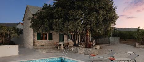 Une piscine avec des chaises longues, un bâtiment en pierre et un grand arbre.