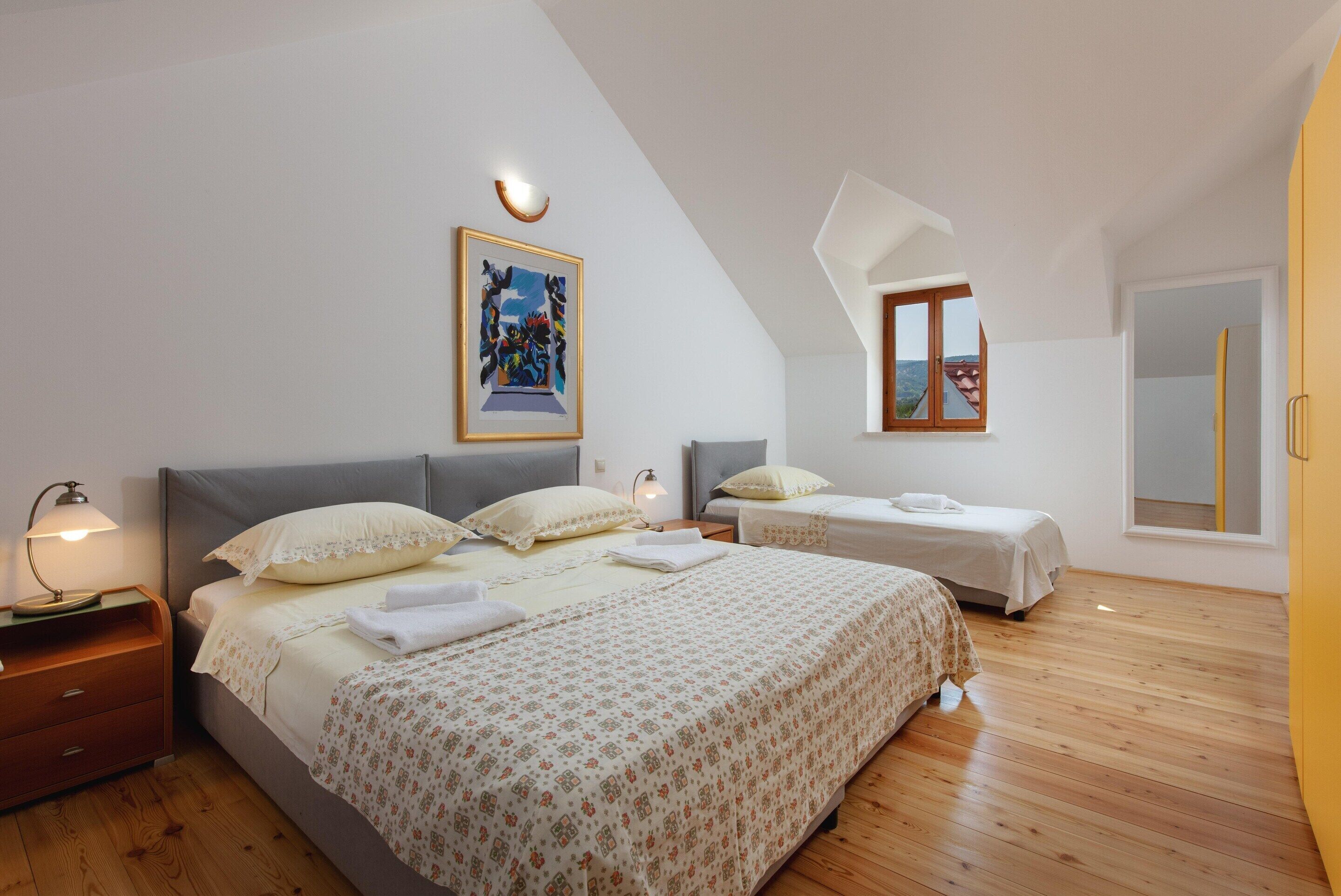 Un dormitorio con una cama grande, dos camas más pequeñas, suelo de madera, un cuadro en la pared y una ventana con vistas.
