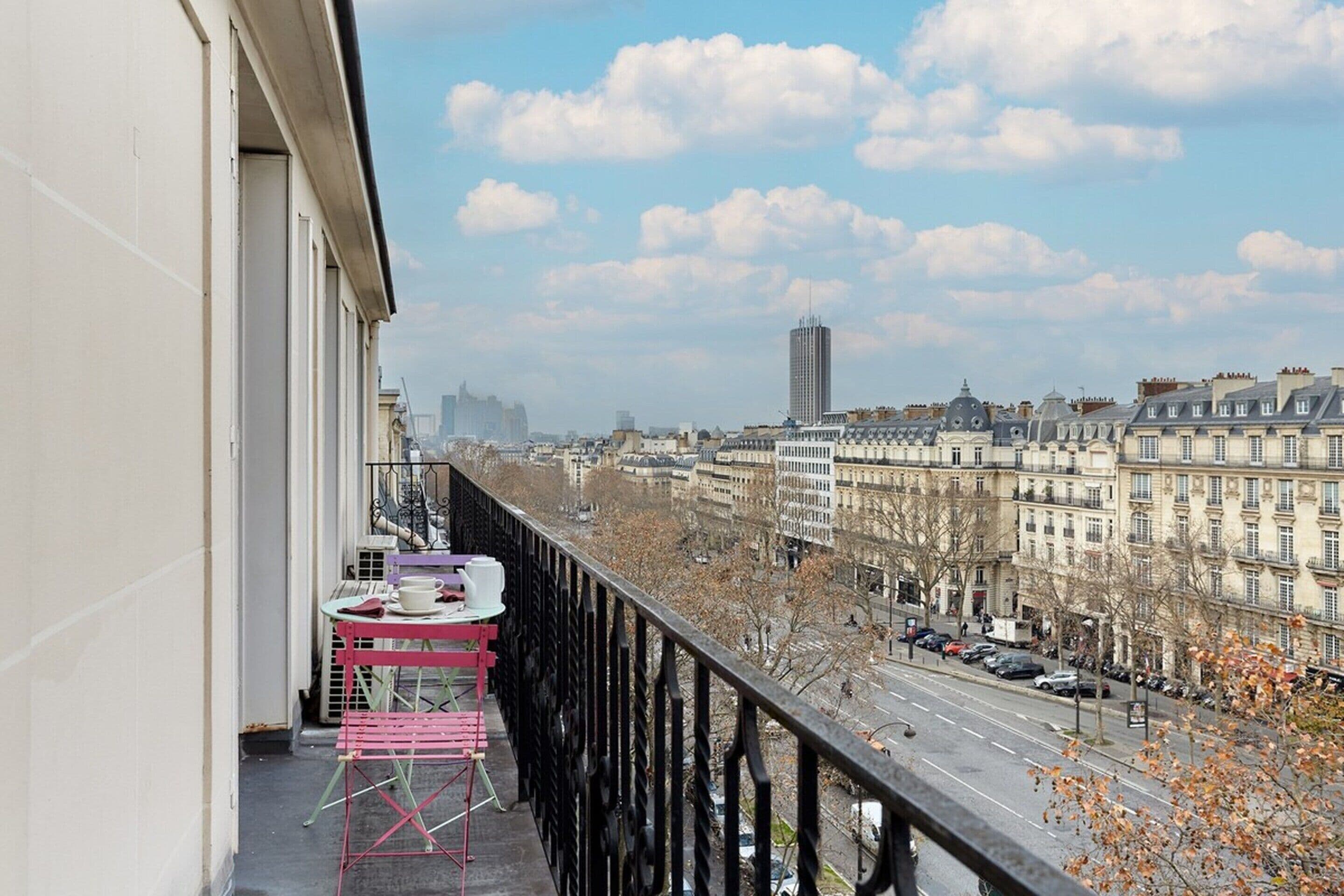 A balcony with a table and chairs, overlooking a city street with buildings and cars.