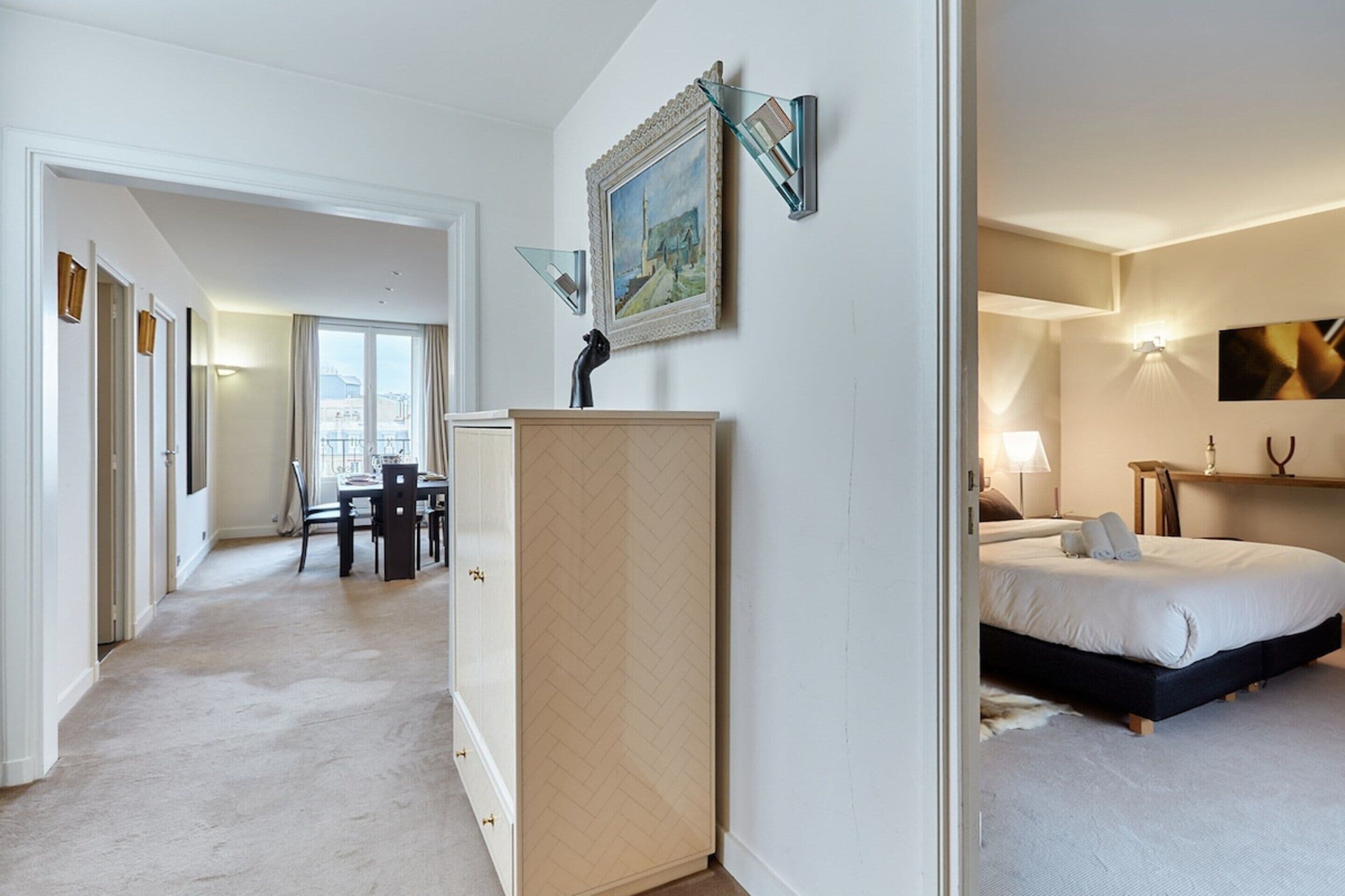 A hotel room with a bed, a desk, and a chair. There is a painting on the wall and a lamp on the bedside table.