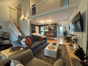 A spacious living area with a grey sofa, a coffee table, and a dining table with chairs. A staircase leads to an upper level with a balcony.