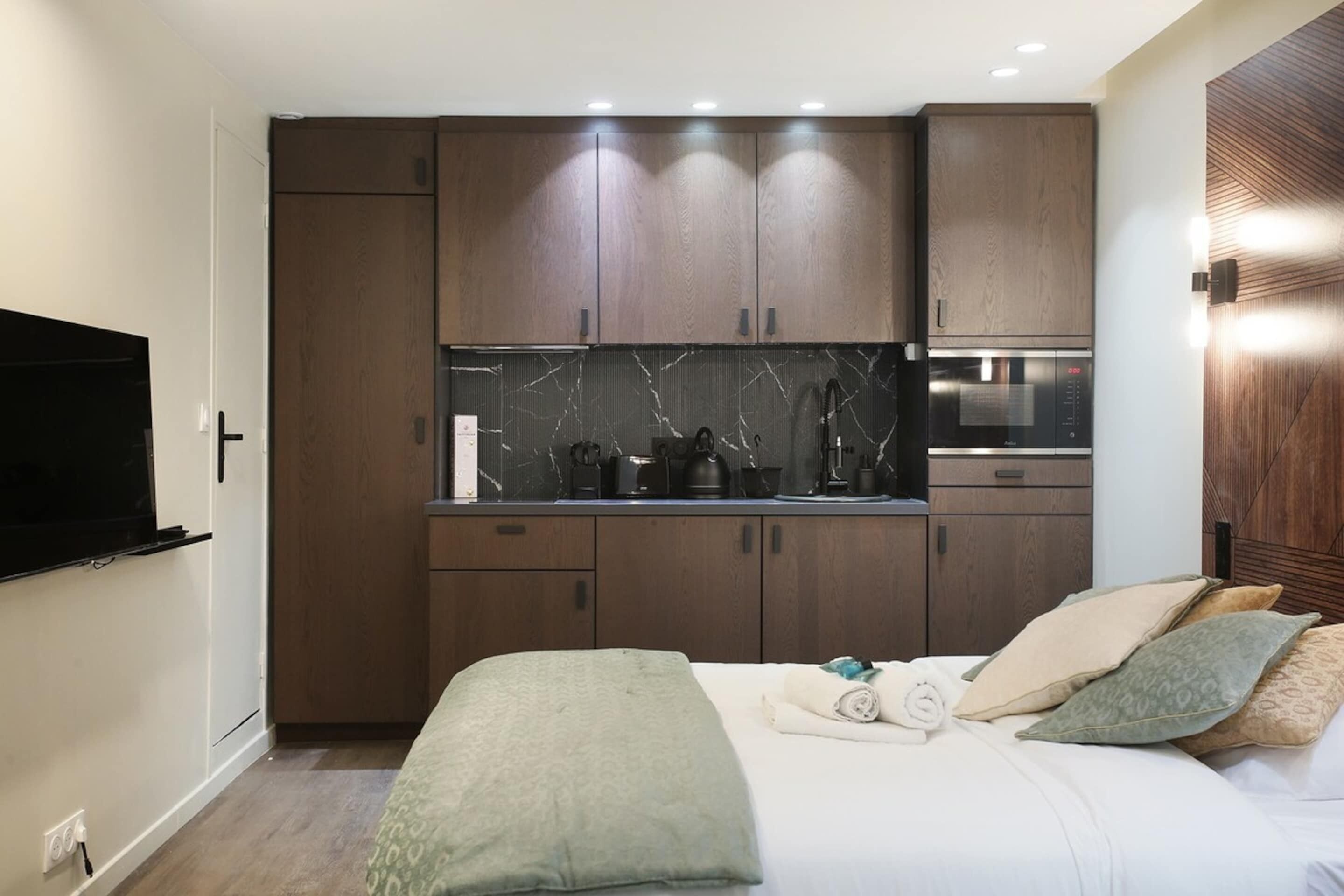 Camera d'albergo compatta con angolo cottura, un letto con cuscini e una TV a schermo piatto montata a parete.