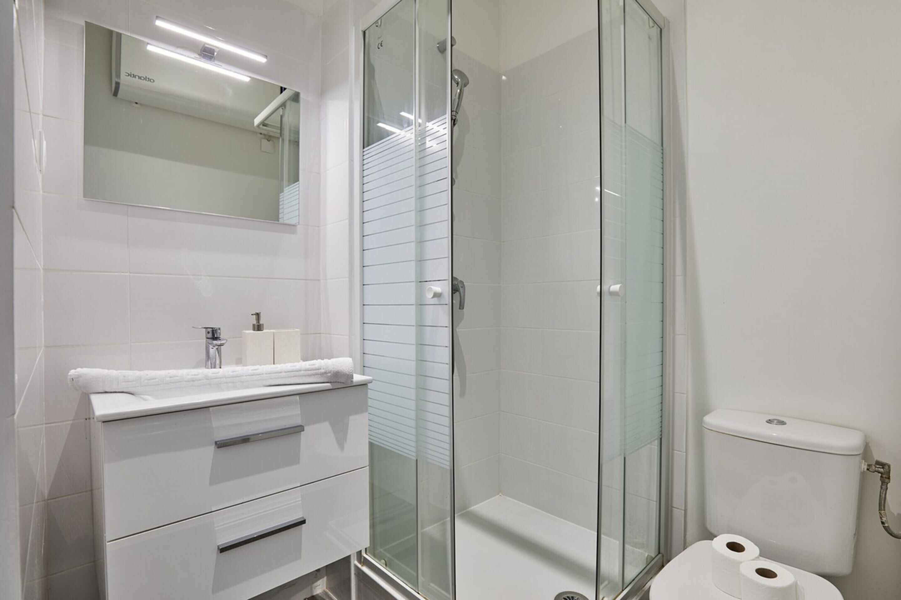 Une salle de bain moderne avec une douche à parois de verre, des carreaux blancs, un meuble-lavabo avec des tiroirs et des toilettes.