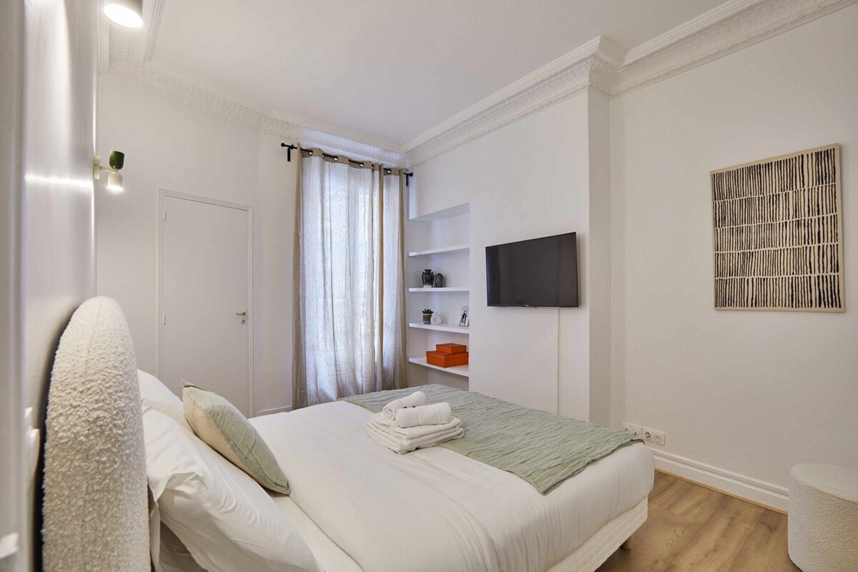 A neatly arranged bedroom with a bed, a television mounted on the wall, and a shelf with decorative items.