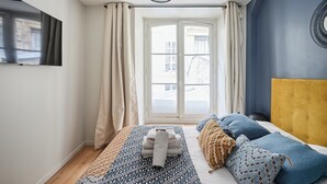 Una camera d'albergo con un letto, una finestra con tende, una TV a parete e un lampadario originale.