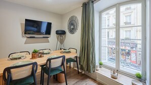 Ein Essbereich mit einem Holztisch, vier Stühlen, einem an der Wand montierten Fernseher und einem Fenster mit Blick auf ein Gebäude.