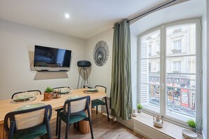 Een eetruimte met een houten tafel, vier stoelen, een aan de muur bevestigde televisie en een raam met uitzicht op een gebouw.