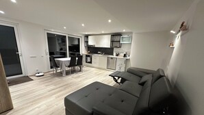 A modern living room with a grey sofa, a dining table with chairs, a kitchen area with white cabinets, and a sliding glass door leading to a balcony.