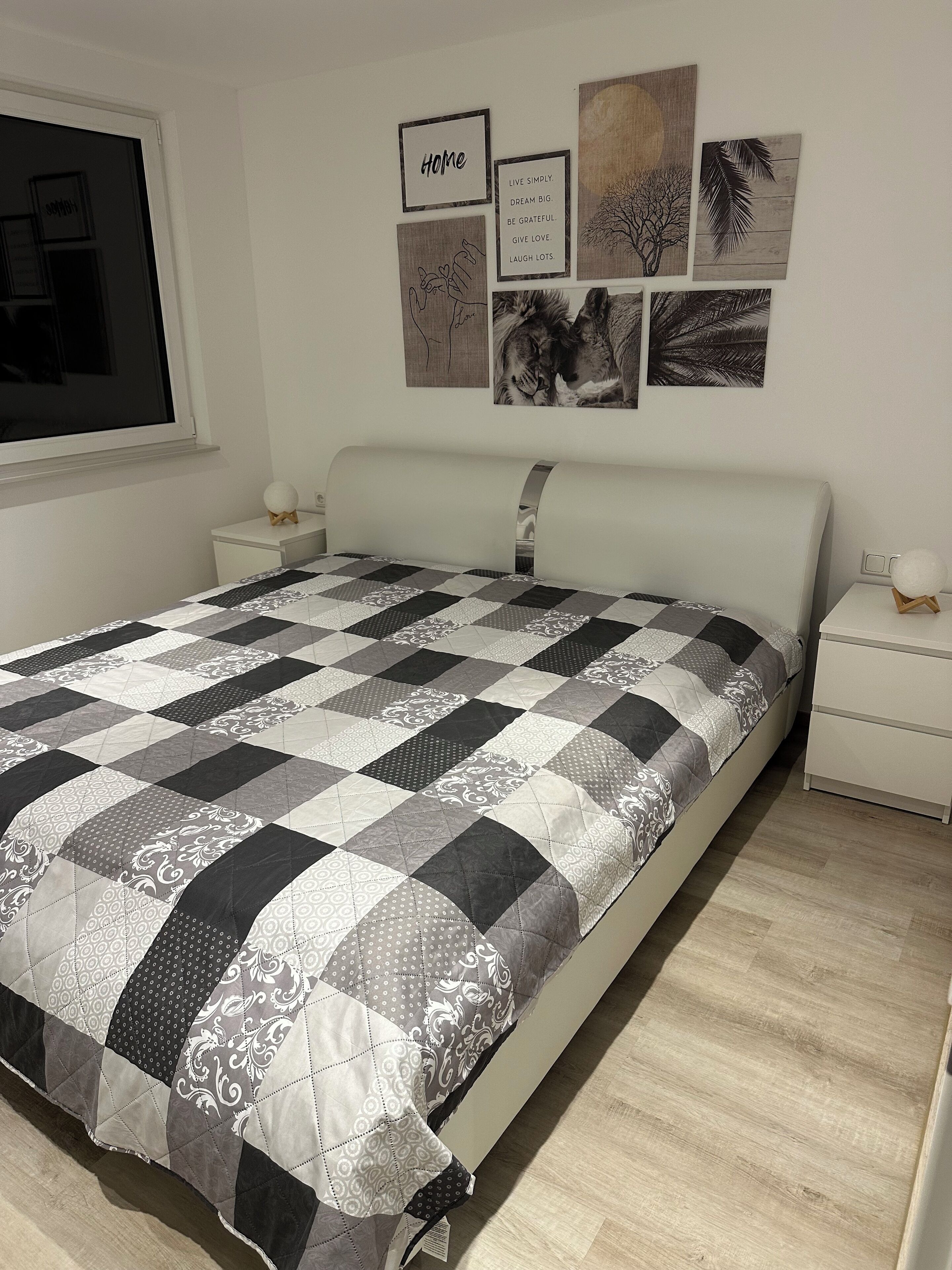A bedroom with a checkered bedspread, a white headboard, and a wall with framed pictures and a window.