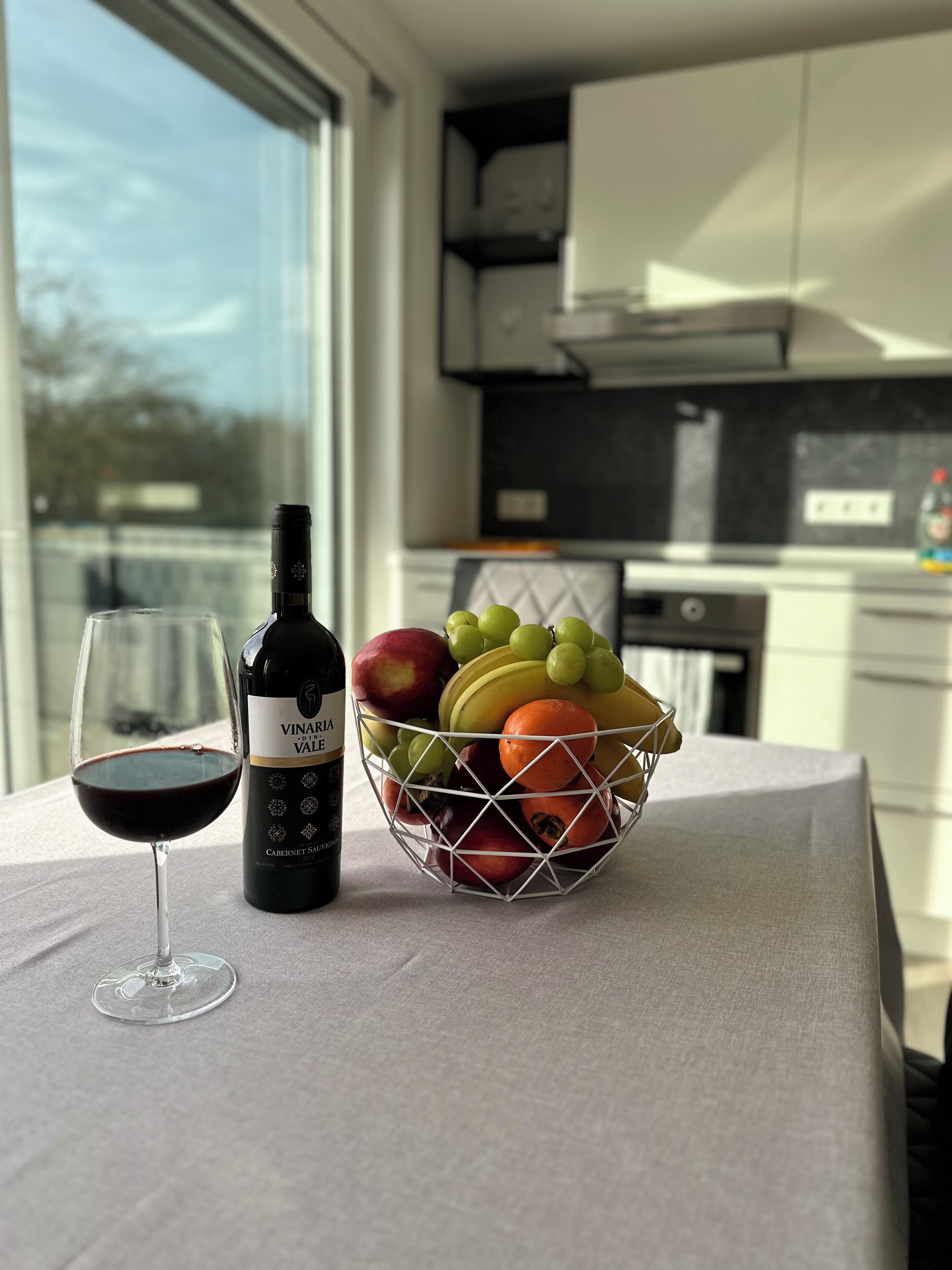 Un tavolo con una bottiglia di vino, un bicchiere di vino rosso e un cesto di frutta contenente uva, mele e arance.