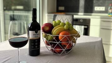 Un tavolo con una bottiglia di vino, un bicchiere di vino rosso e un cesto di frutta contenente uva, mele e arance.