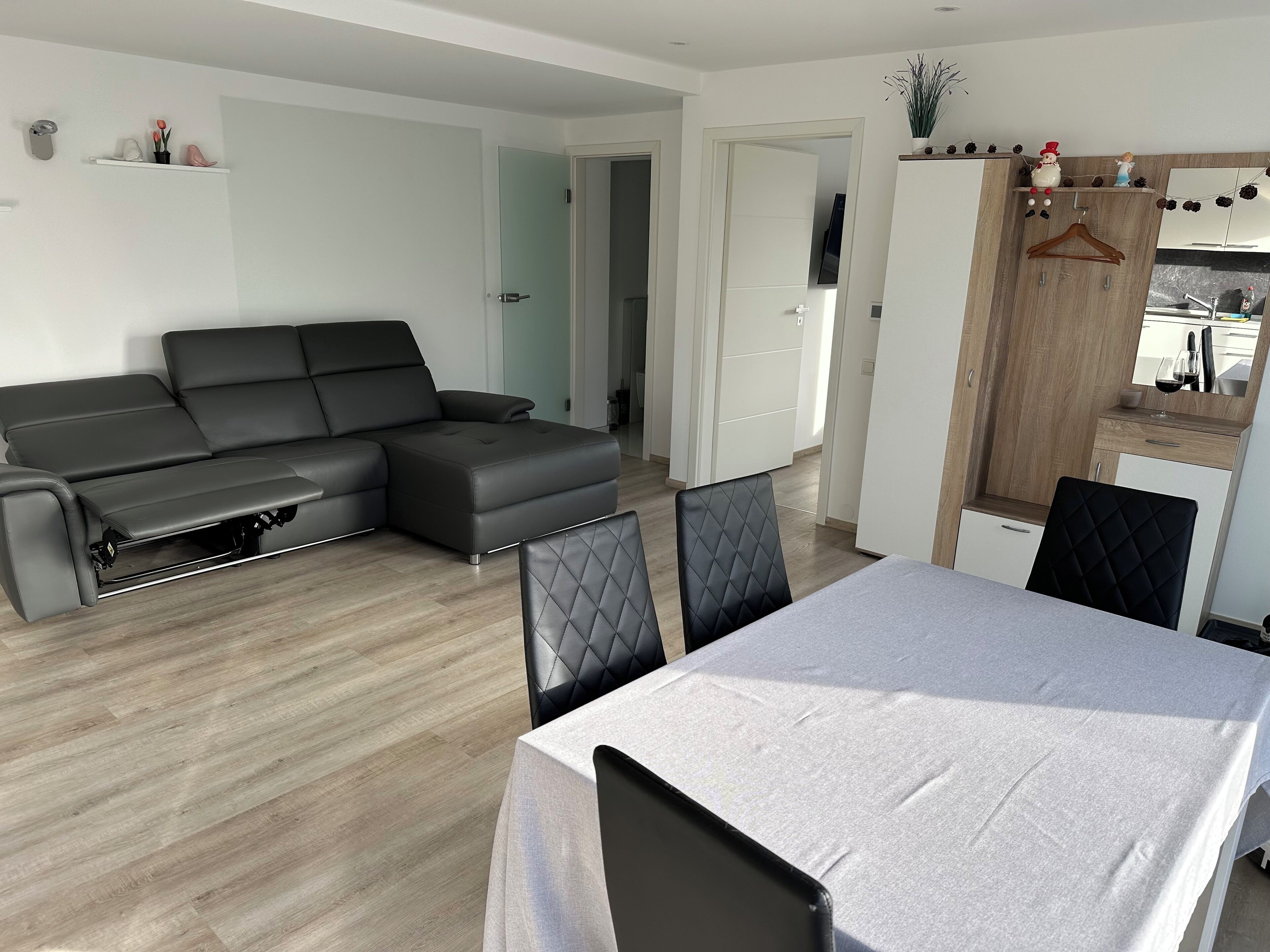 Un salon moderne avec un canapé d’angle, une table à manger avec des chaises et un coin cuisine doté d’un meuble en bois et d’une plante suspendue.